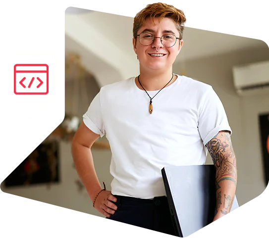 Person with glasses and tattoos holding a laptop, smiling, wearing a white shirt and standing indoors.