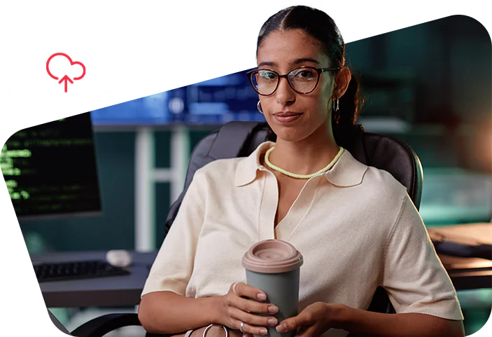Woman with glasses holding a coffee cup, seated at a desk with multiple computer monitors.