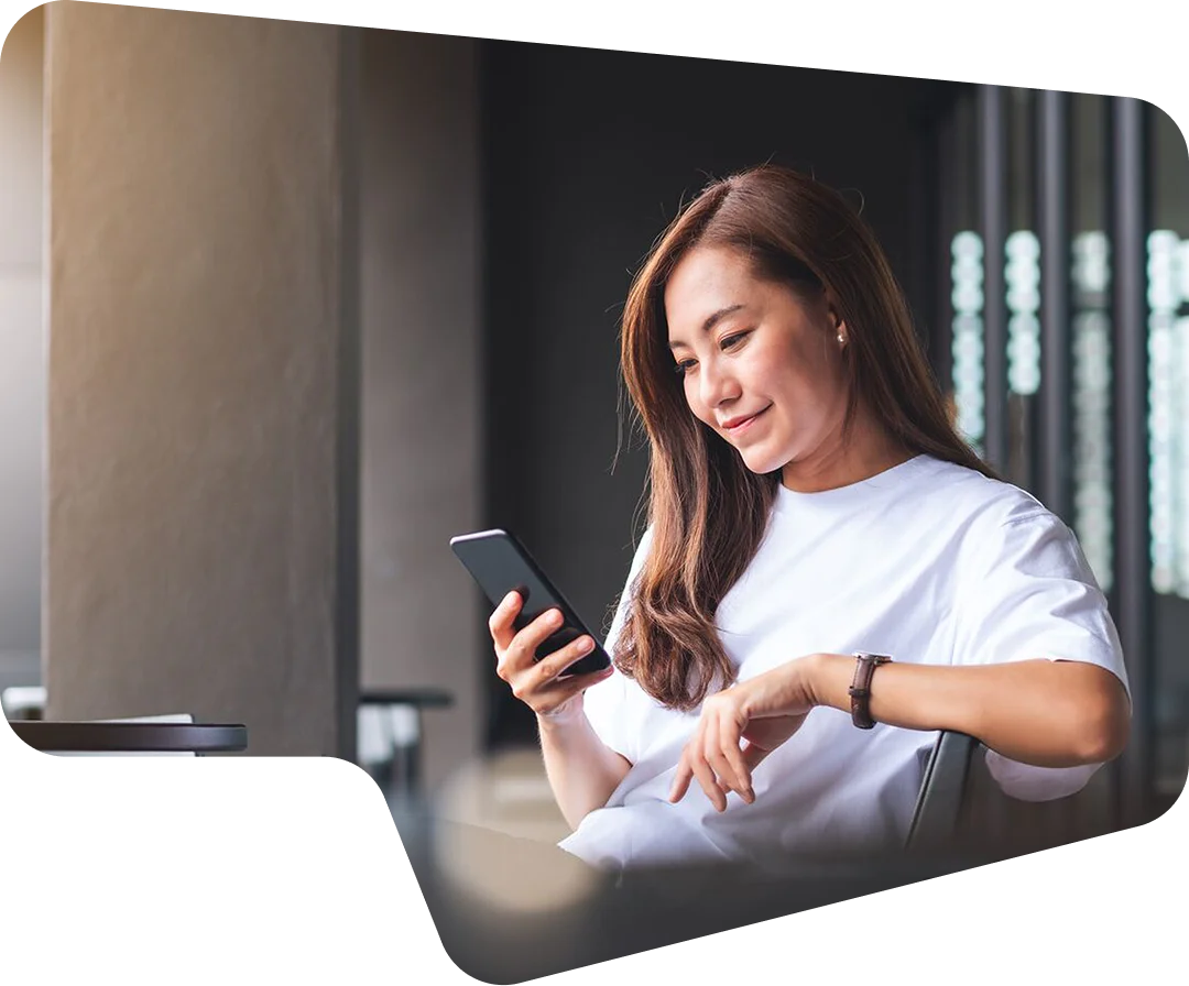 A woman in a white shirt sitting and smiling while looking at her smartphone.