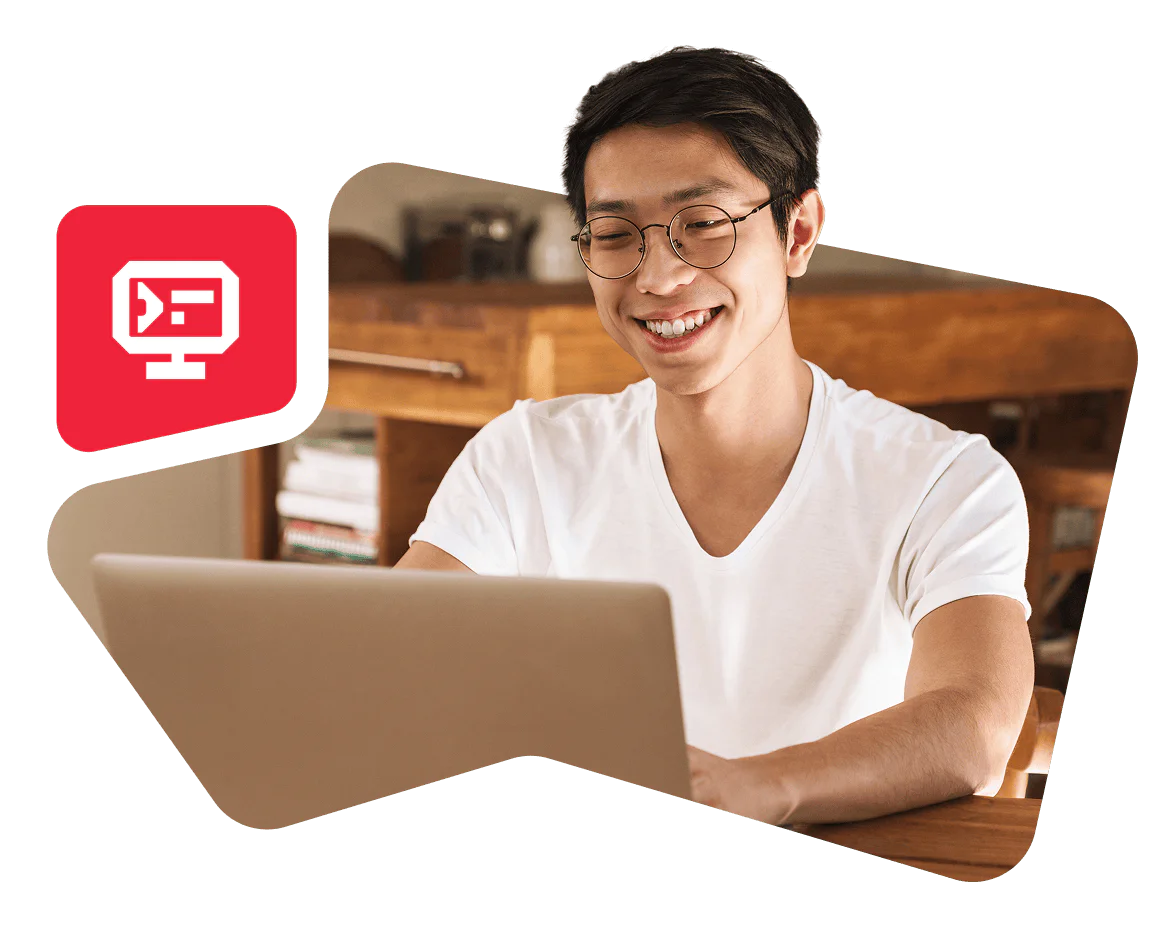 A man wearing glasses and a white t-shirt is smiling while using a laptop at a wooden desk.