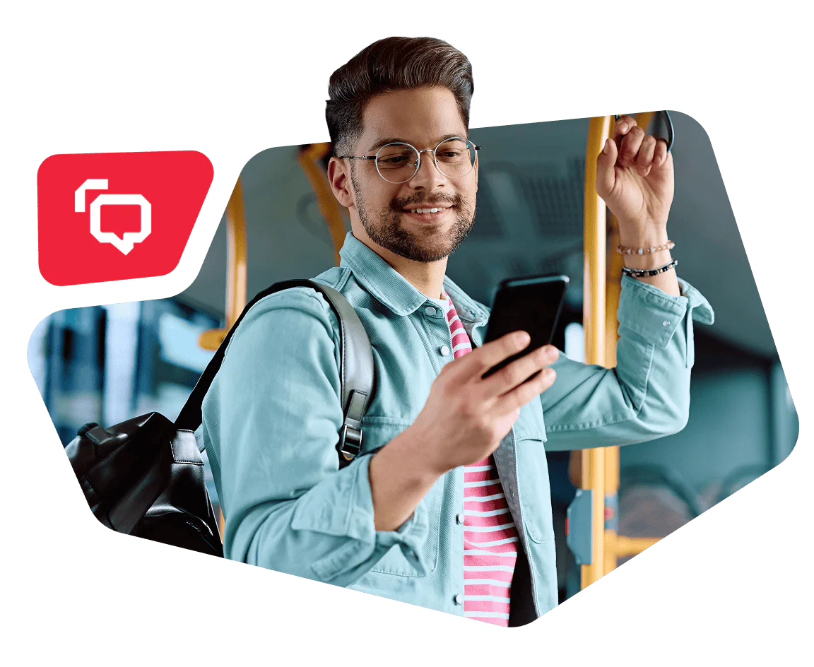 Man smiling while looking at his smartphone on a bus, holding onto a handrail and carrying a backpack.