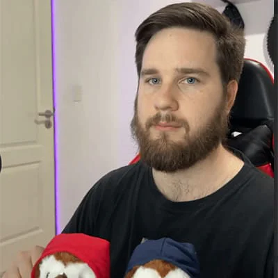 Bearded man holding two stuffed animals dressed in red and blue hoodies.