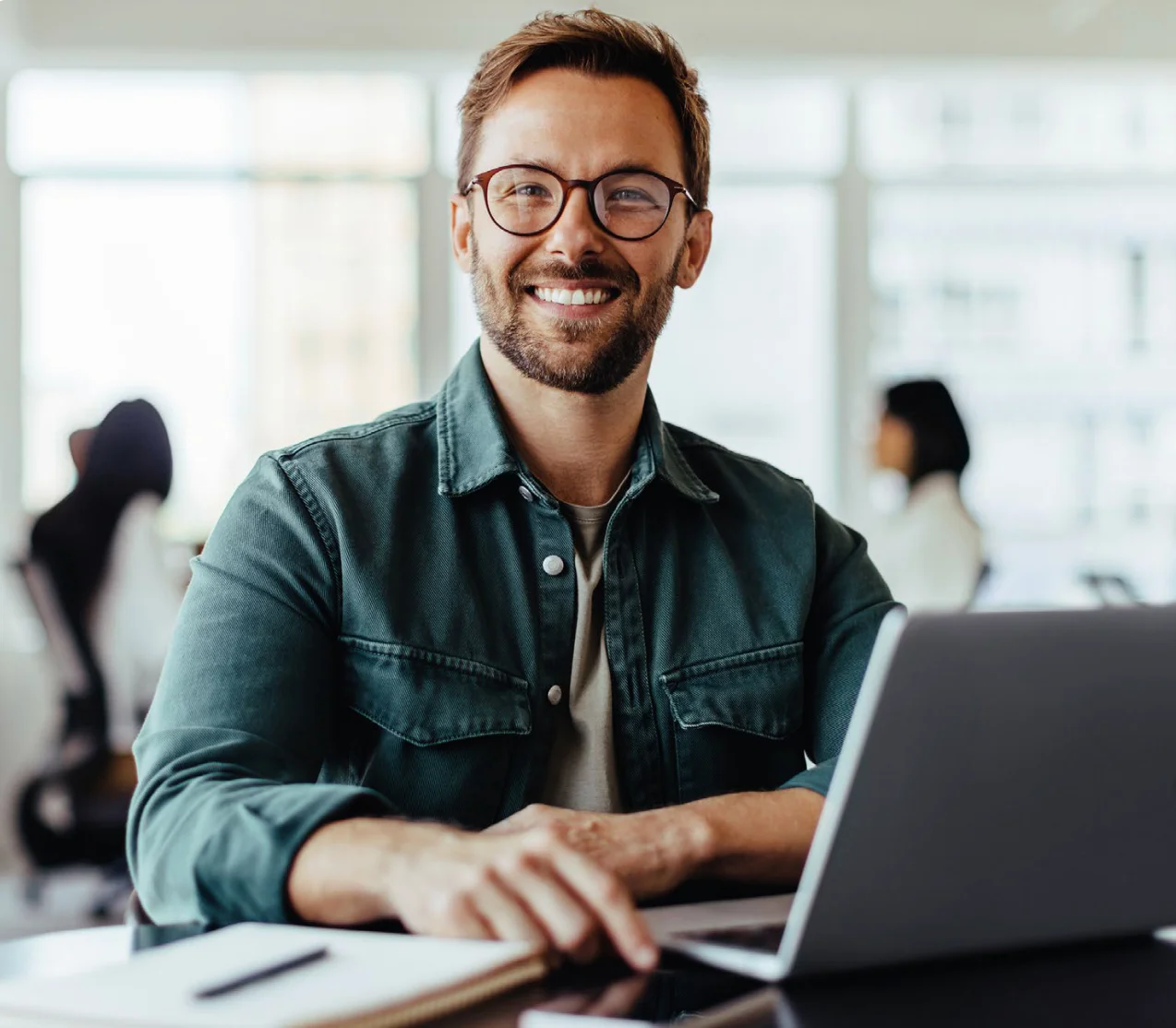 Man with glasses working on a laptop and smiling in a bright office setting.