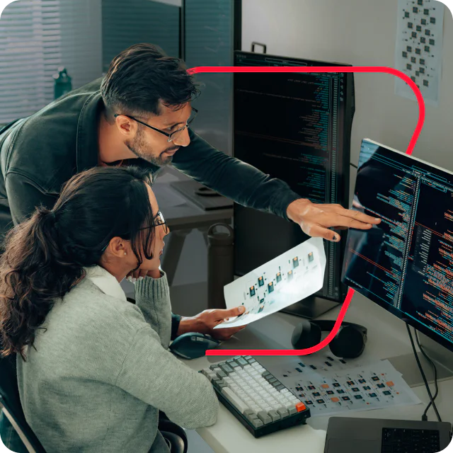 Two people reviewing code and diagrams on multiple computer monitors in an office.