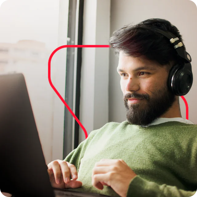 A man with headphones and a green sweater using a laptop near a window.