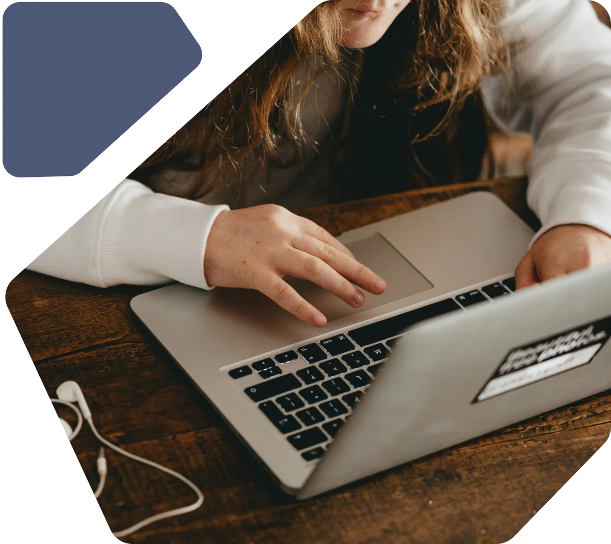 Person in white shirt using a laptop on a wooden table with earphones nearby.