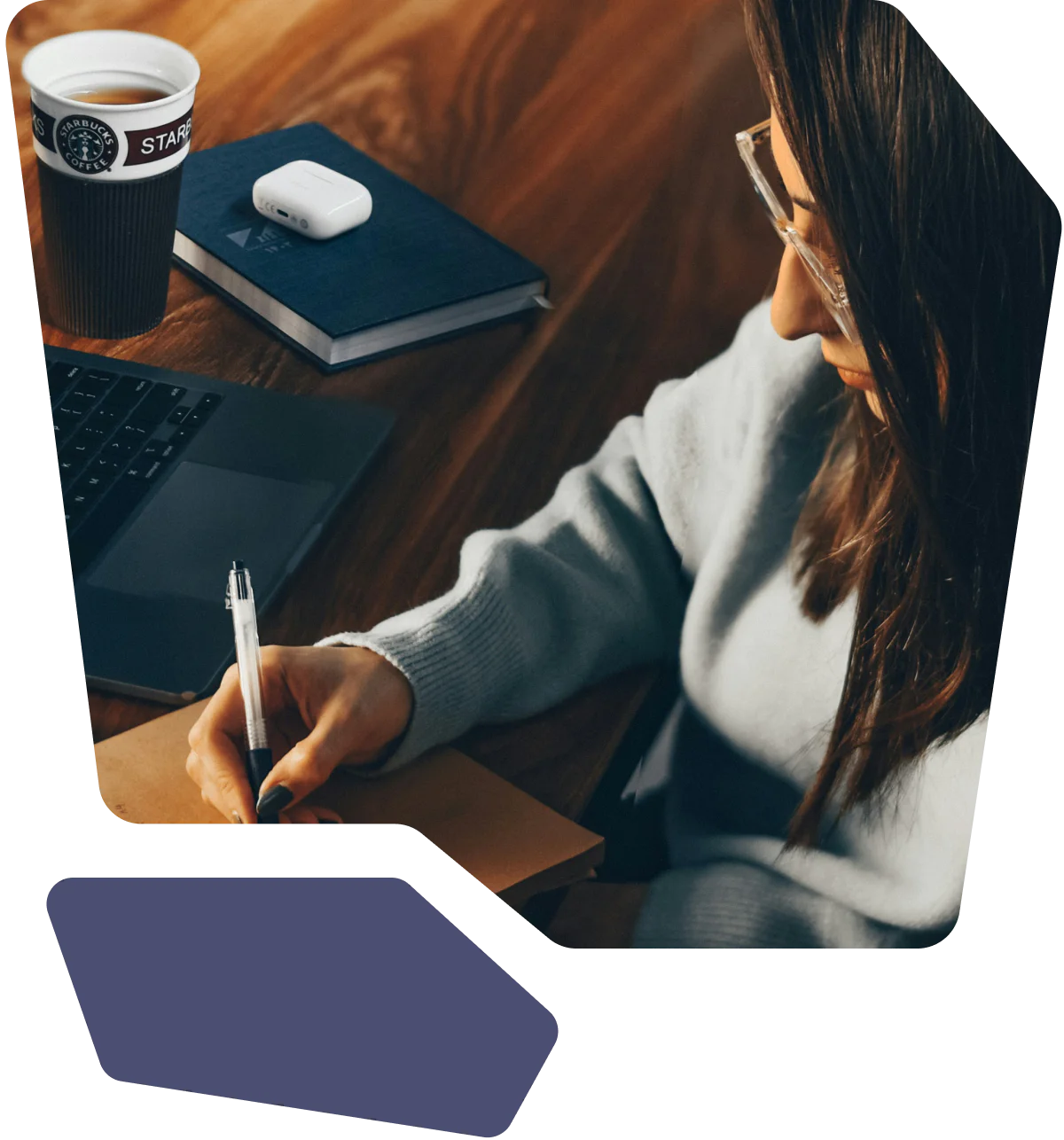 Person writing in a notebook with a laptop, coffee cup, and book on a wooden table.