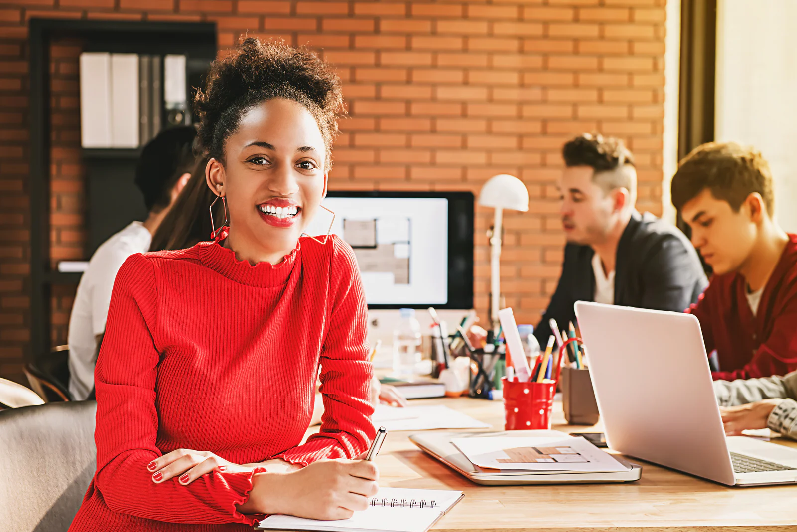 Woman in red sweater smiling at a desk with colleagues working in the background.
