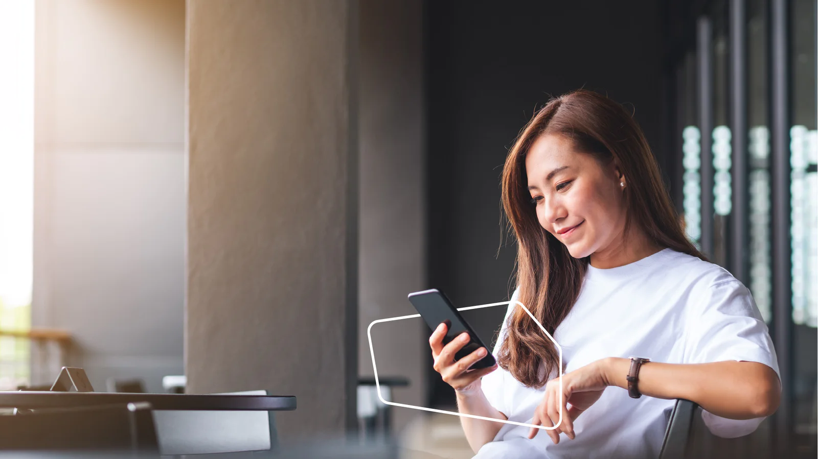 A young woman in a white shirt looks at her smartphone while sitting at a cafe.