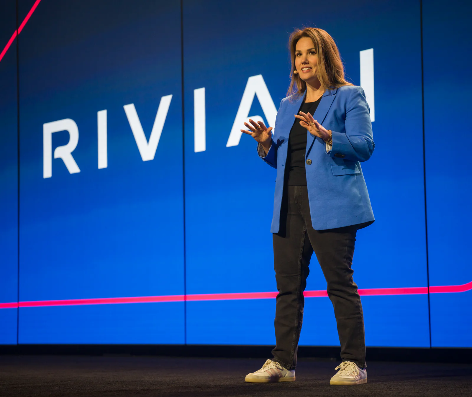 Woman in a blue blazer speaking on a stage with a large Rivian logo in the background.