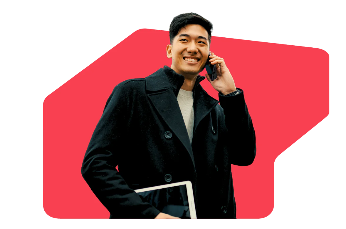 Man in a black coat smiling while talking on the phone, holding a tablet against a red background.