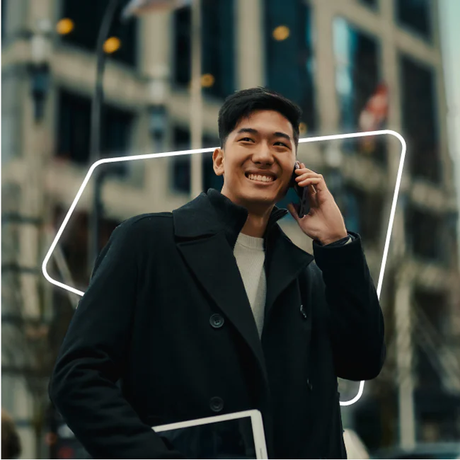 Man in black coat smiling while talking on the phone, holding a tablet, with a city building in the background.