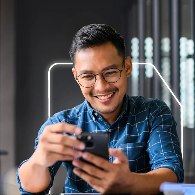 Man in glasses and blue checkered shirt looking at smartphone and smiling in an office.