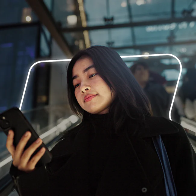 Woman in black coat using smartphone on escalator with neon light frame behind her.