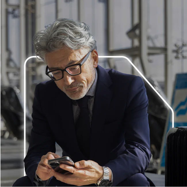 Elderly businessman with glasses using smartphone, suitcases in the background.