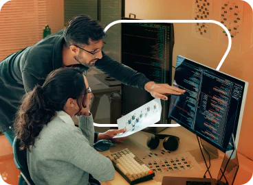 A man and a woman working together on a computer, reviewing code and charts on a monitor and paper.
