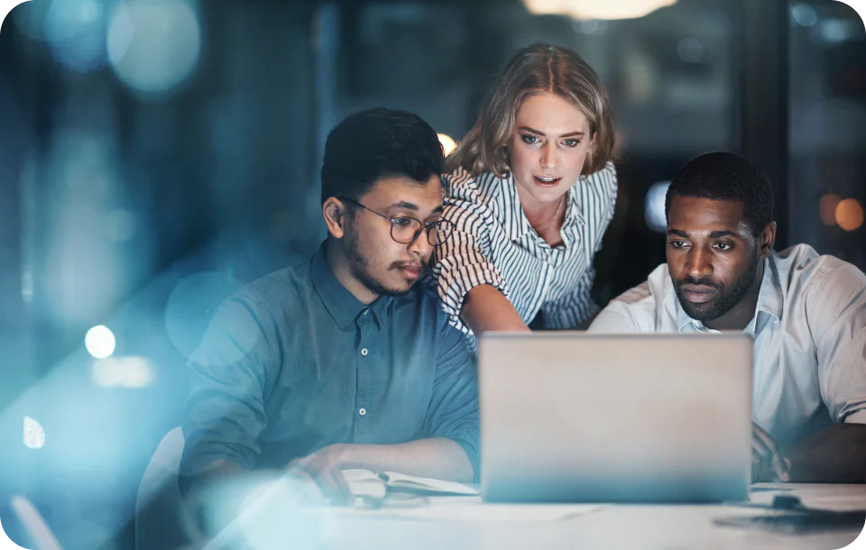 Three colleagues working together on a laptop at a desk in an office environment at night.
