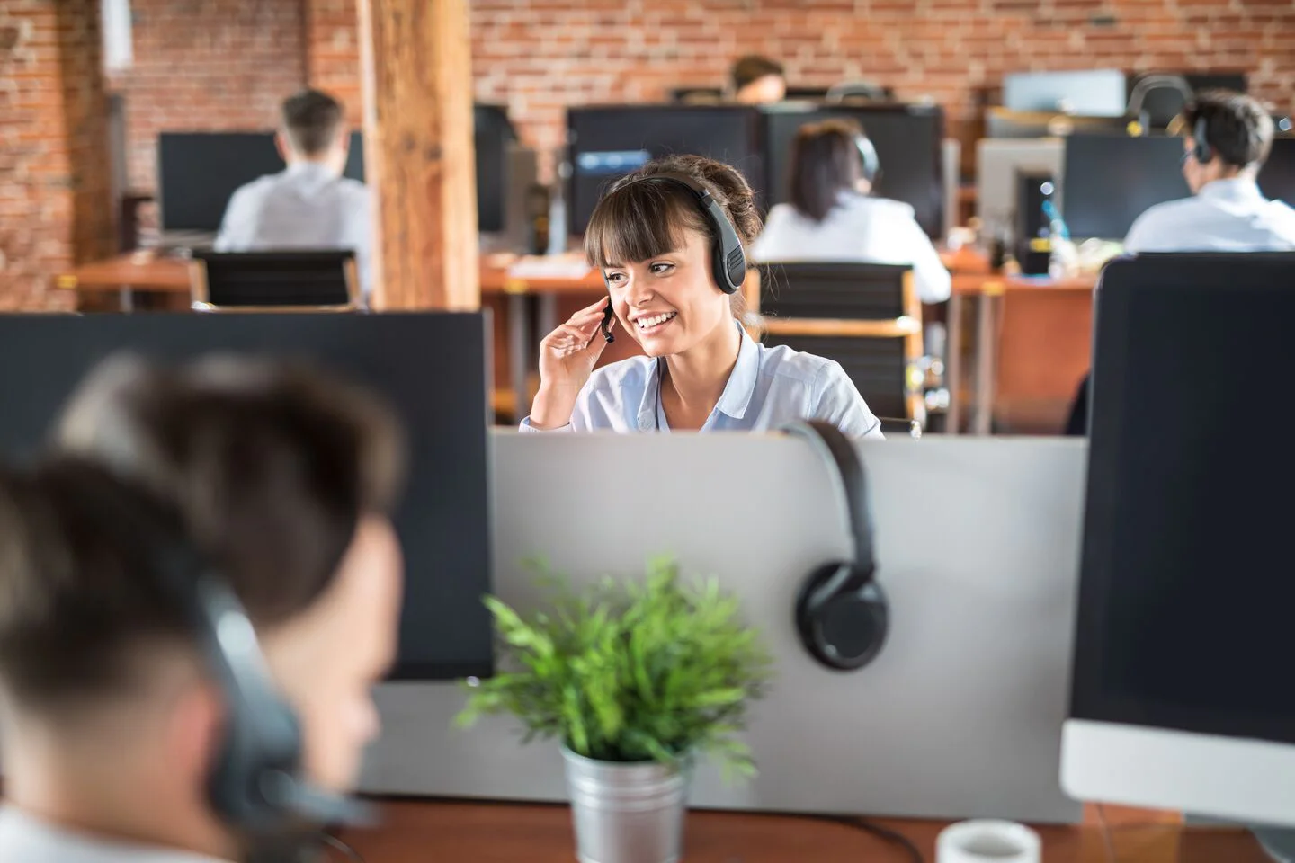 Call center agents wearing headsets, working at desks with computers in a brick-walled open office.