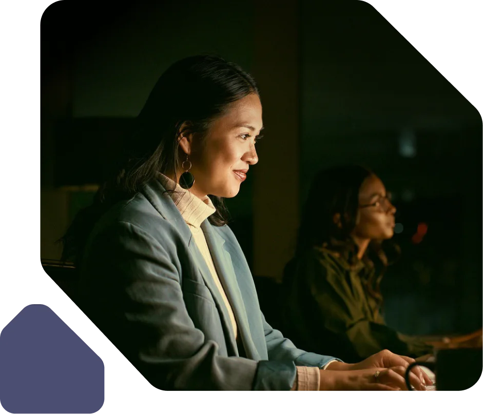 Two women working intently on computers in a dimly lit office setting.