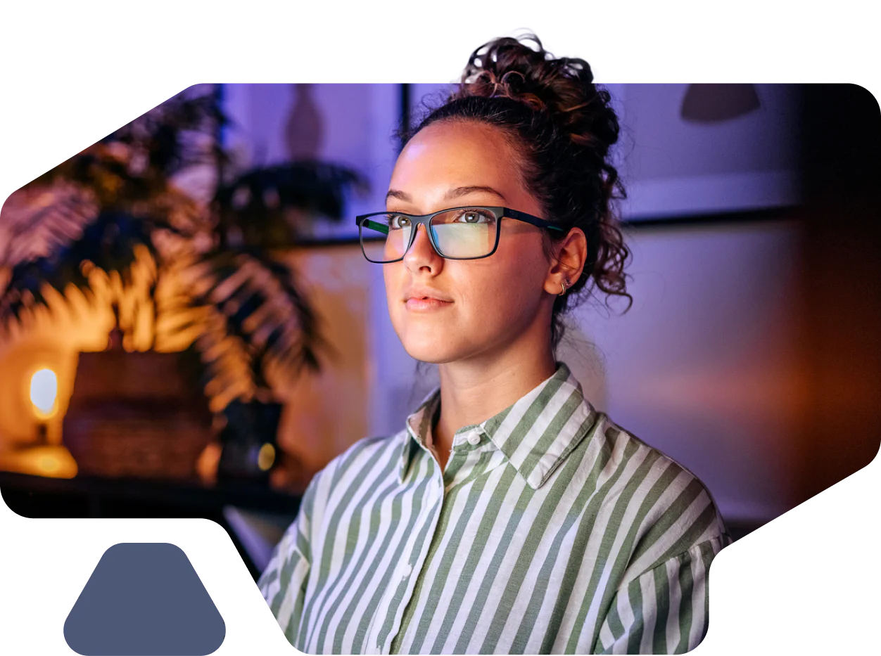 Woman with glasses and curly hair working at her computer in a modern office with warm lighting.