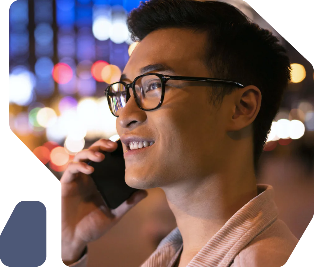 Man with glasses smiling while talking on a smartphone at night with blurred city lights in the background.