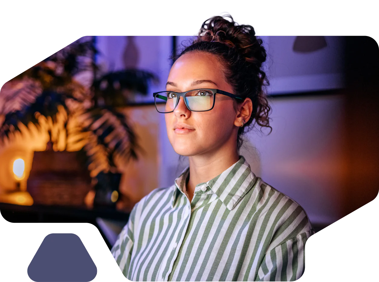 A woman with glasses, wearing a striped shirt, is looking ahead in a dimly-lit room with indoor plants.