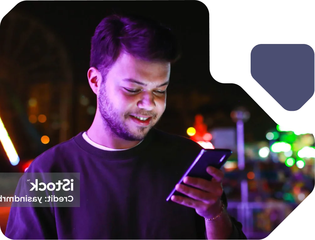 Young man smiling while using smartphone at night. Man using phone, standing outside with colorful lights in the background at nighttime.