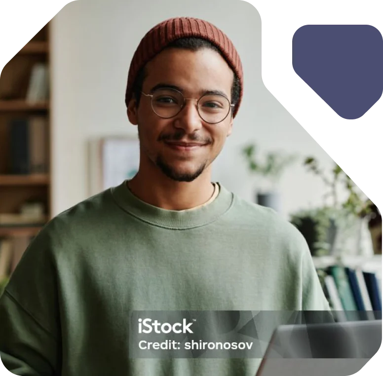 Young Man Smiling While Holding a Tablet A young man in glasses and a beanie smiles while holding a tablet in a cozy room.