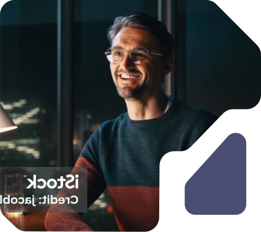 Man smiling while working late at night with city lights in the background. Man with glasses and a sweater smiling at a desk with city lights visible through the window.