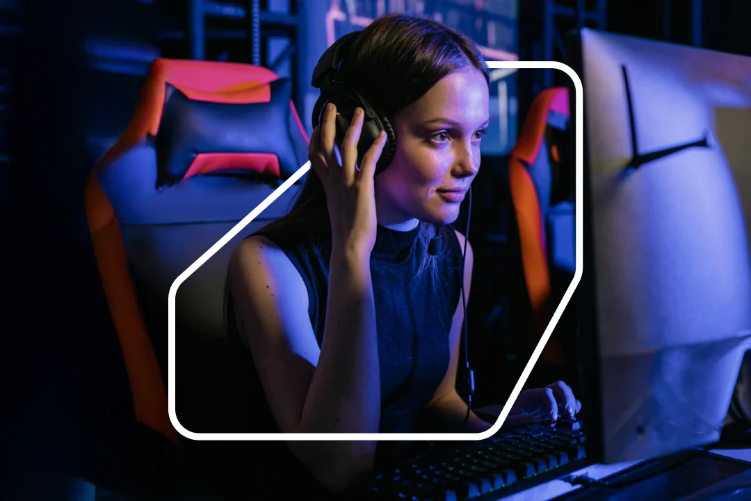 Woman wearing headphones and gaming at a computer in a dimly lit gaming station.