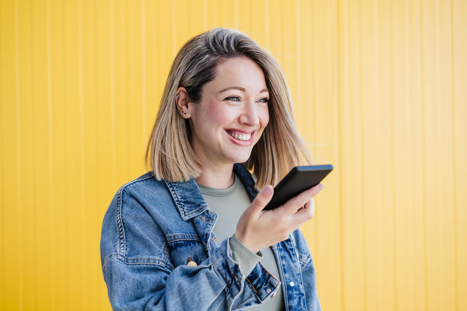 Woman Smiling While Using Smartphone Woman in denim jacket smiling and using a smartphone against a bright yellow background.