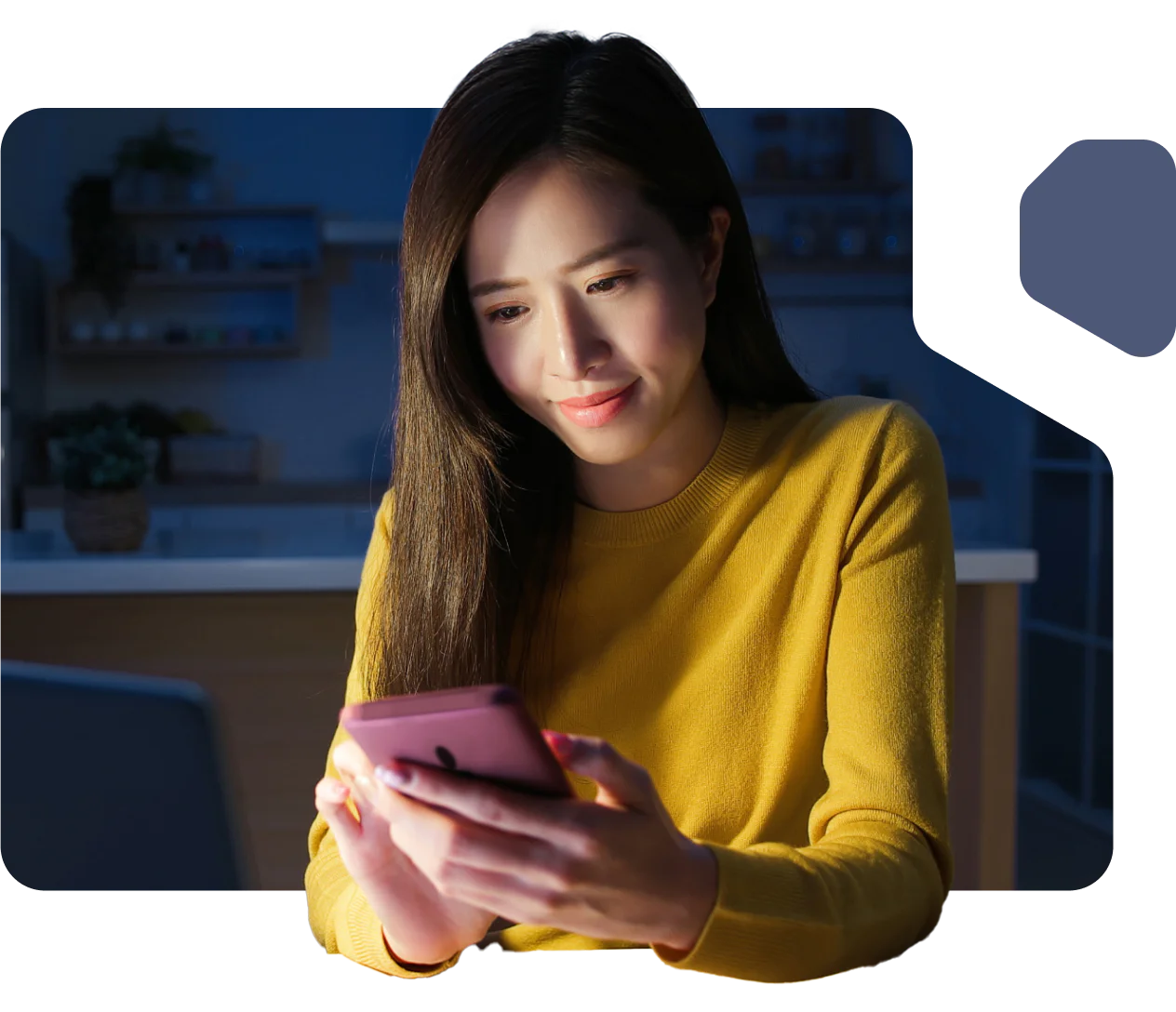 A woman in a yellow sweater looks at her smartphone, sitting in a warmly lit kitchen.