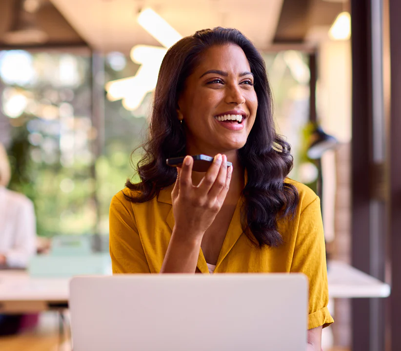 Woman in a yellow shirt smiles while speaking into a smartphone, sitting at a desk with a laptop