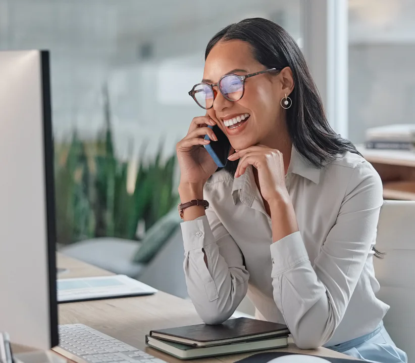 Smiling woman with glasses talking on the phone and working on a computer at her desk.