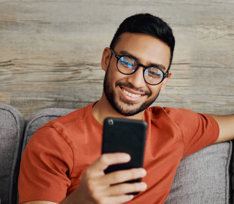 Man with glasses smiling while looking at a smartphone, sitting on a gray couch in a relaxed pose.