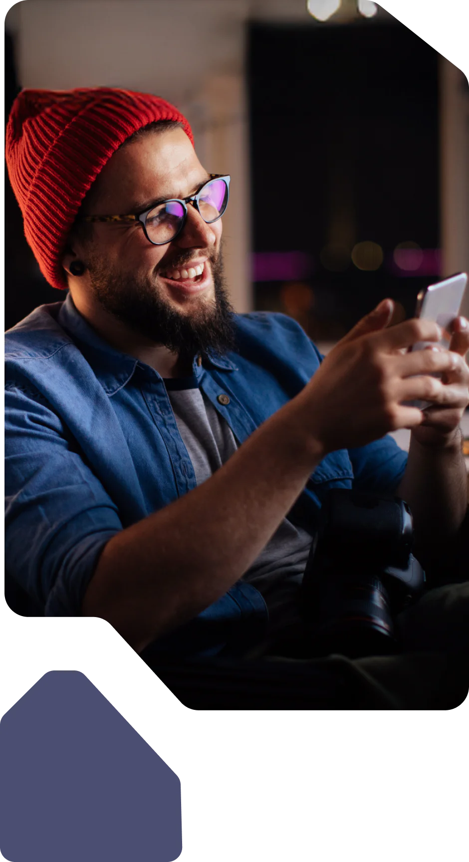 Man with glasses and a red beanie, smiling and using a smartphone in a cozy setting.