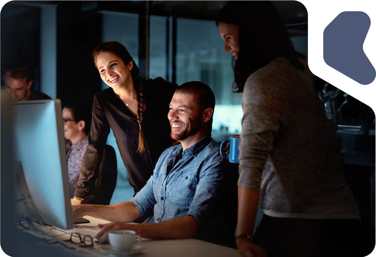 A group of colleagues smiling and working on a computer together in an office setting at night.