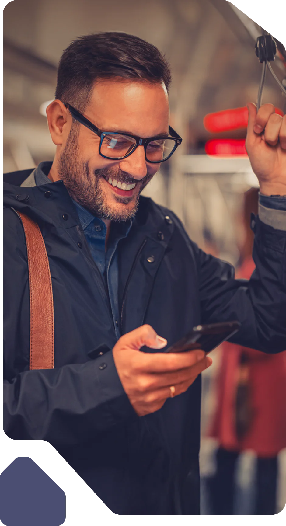 Man wearing glasses and a dark jacket smiles while looking at smartphone on public transport.