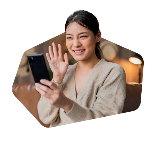 A woman in a beige sweater smiling and waving at her smartphone.