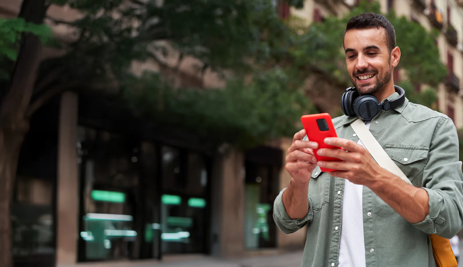 Young caucasian man holding mobile on the street in the city. Happy male wearing headphones and using cell outdoors, looking and checking his social media. Vertical image with copy space.