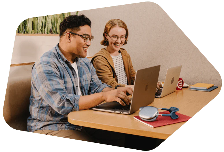Two people smiling and working on laptops at a shared desk with notebooks and headphones.