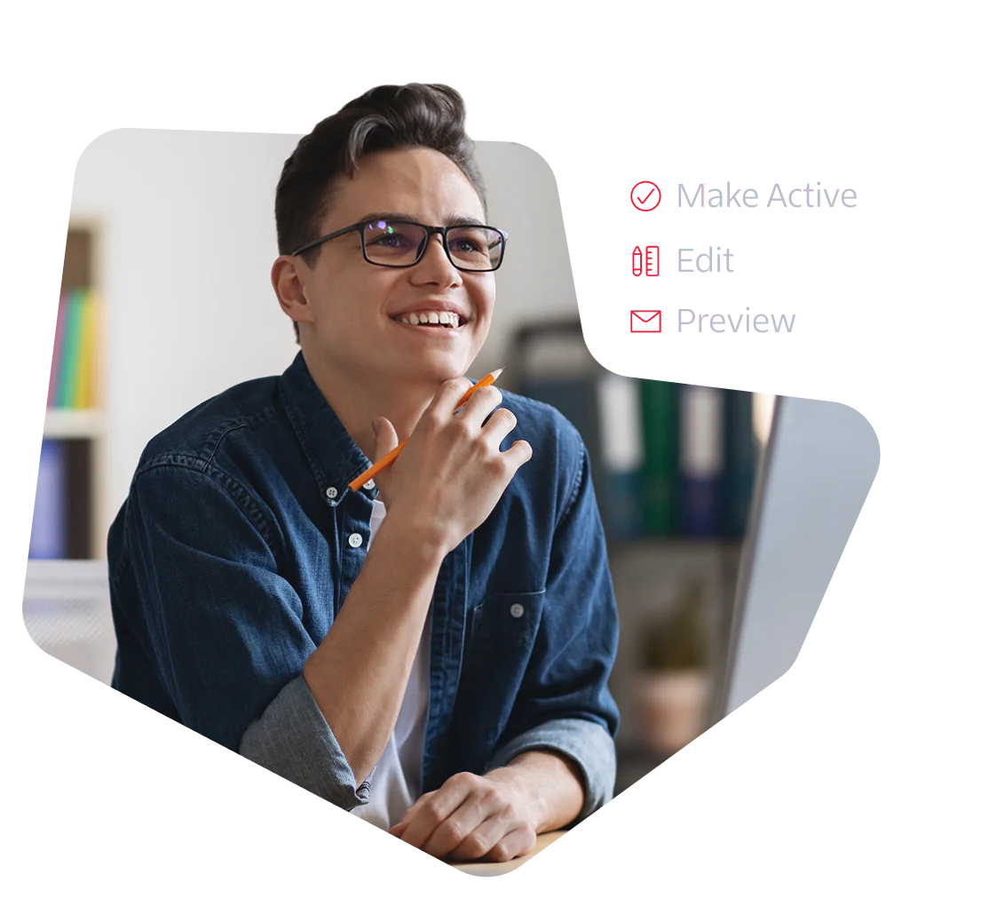 A young man with glasses and a denim shirt smiles while working at a computer.