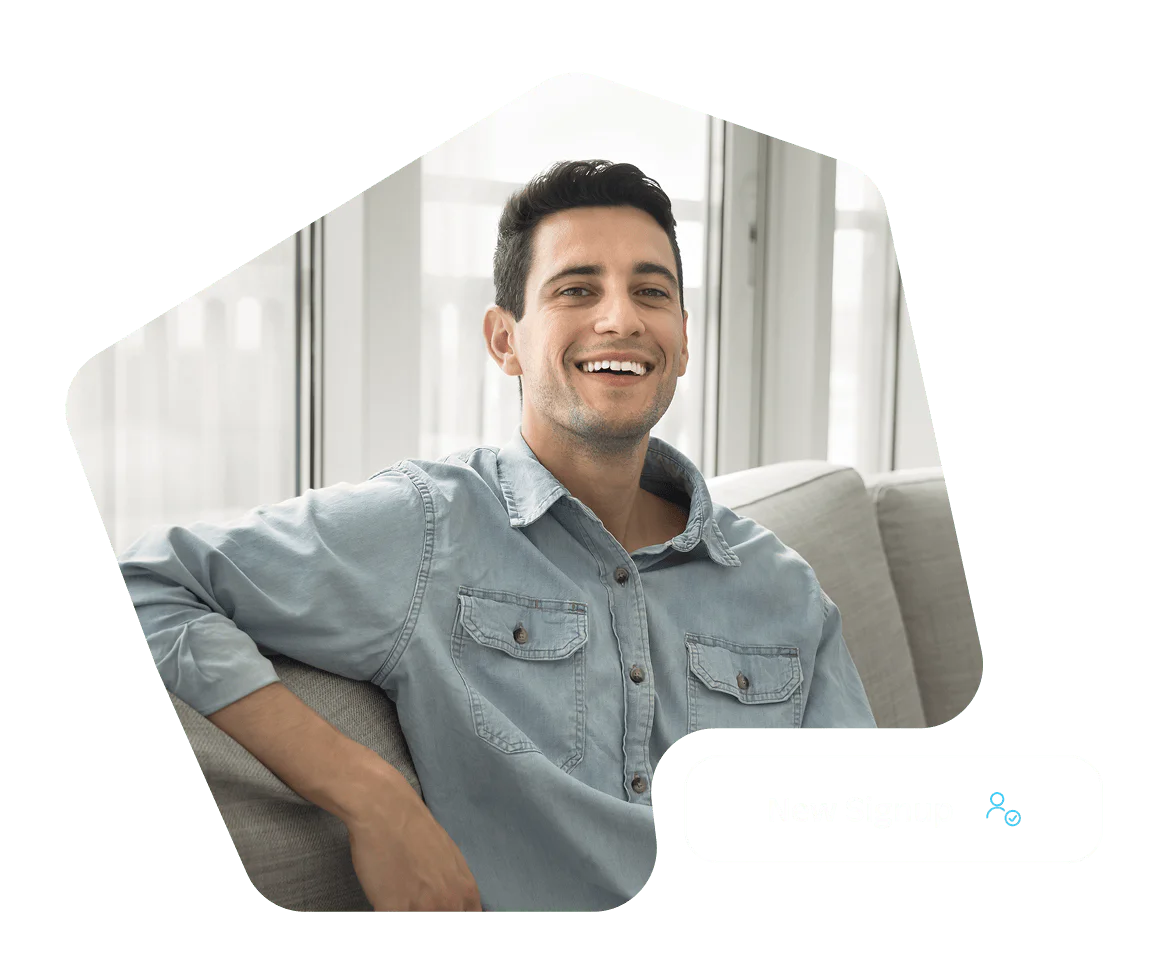 Smiling man in a light blue denim shirt sits on a gray couch in a bright room.