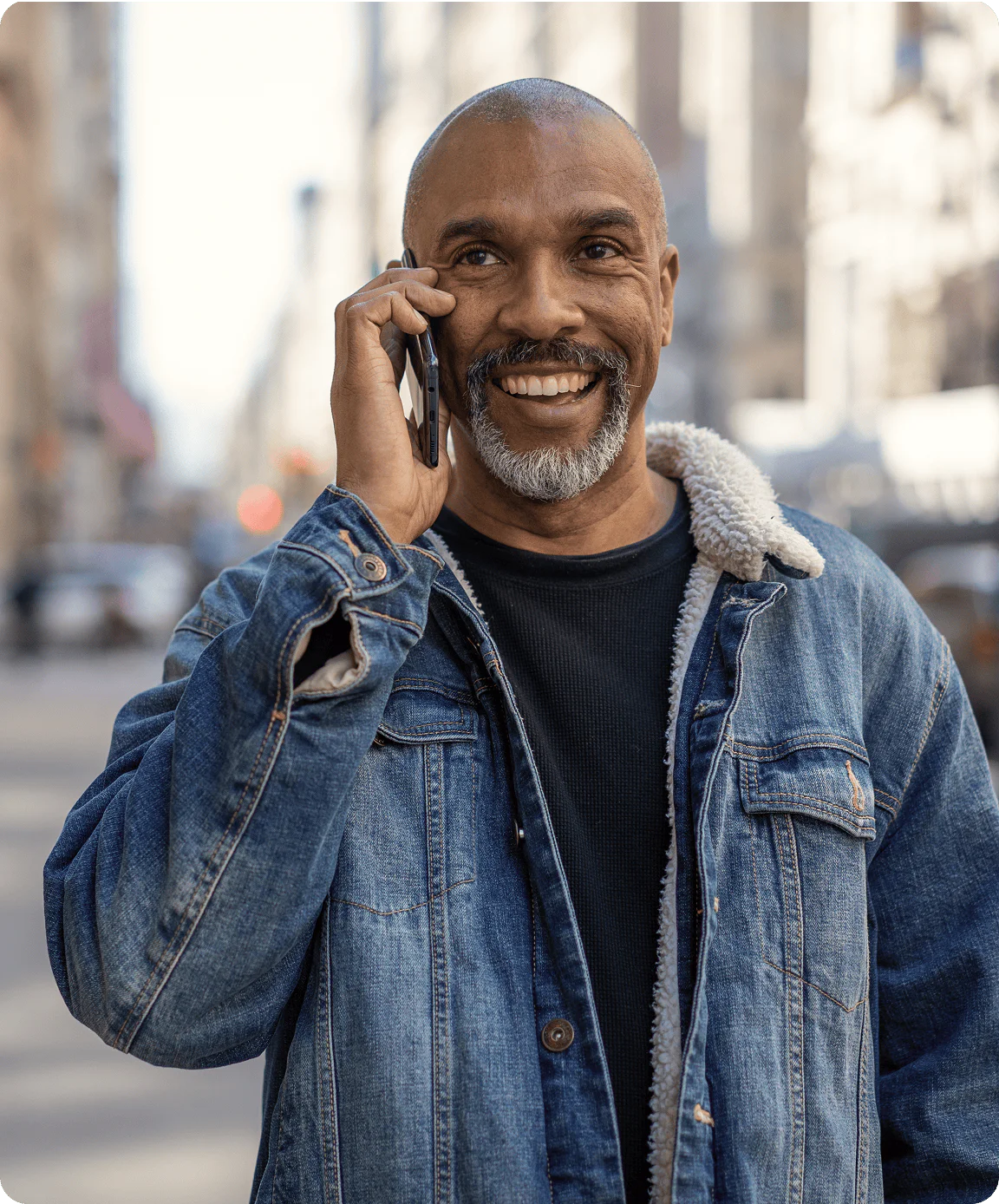 Smiling man in denim jacket uses phone outdoors in urban area.