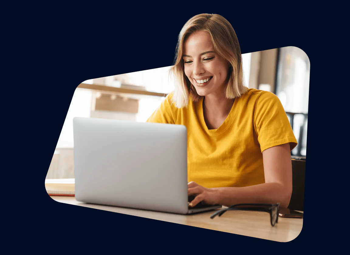 Smiling woman in yellow shirt using a laptop at a wooden desk in a modern workspace