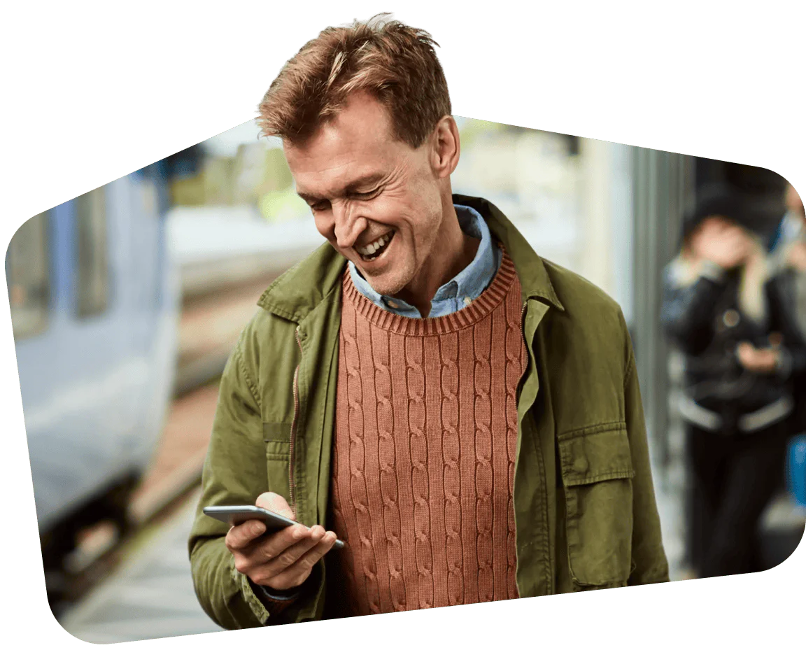 Man in green jacket and orange sweater smiling at his smartphone on a train platform.