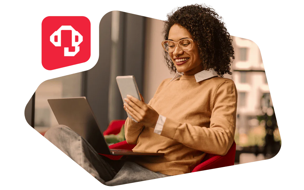 Smiling Woman Using Smartphone and Laptop in Modern Setting Woman with curly hair in glasses using a smartphone and laptop, smiling in a cozy indoor setting.