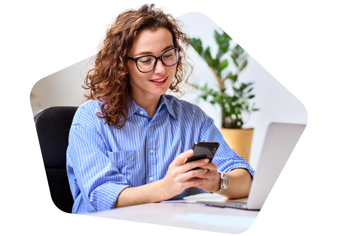 Woman in blue shirt and glasses using smartphone at a desk with a laptop.