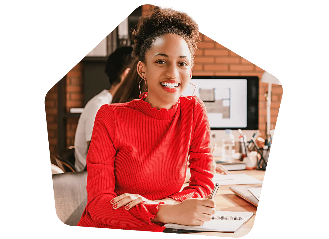 Woman in red sweater with a notebook, smiling at desk in office environment.