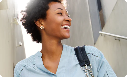 Woman with curly hair and flower earrings smiling while standing outdoors wearing a blue top.